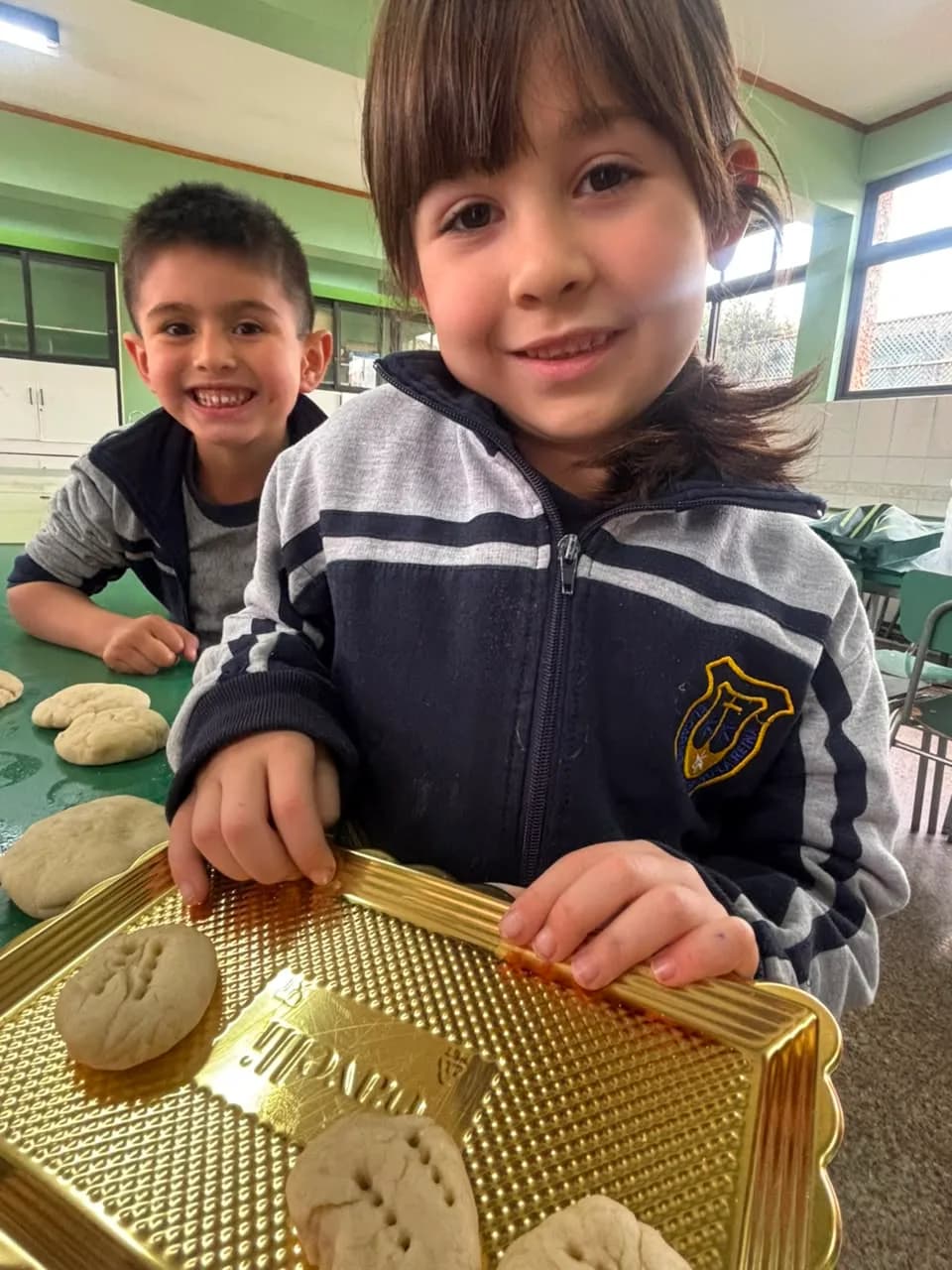 Dos párvulos sonríen a cámara; uno sostiene una bandeja con masas y lleva el polar con el escudo del colegio.