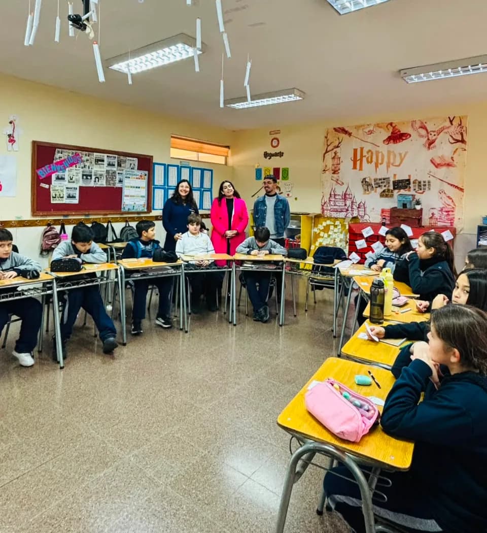 Tres adultos acompañan a un curso en la sala, de pie entre los pupitres durante una actividad.