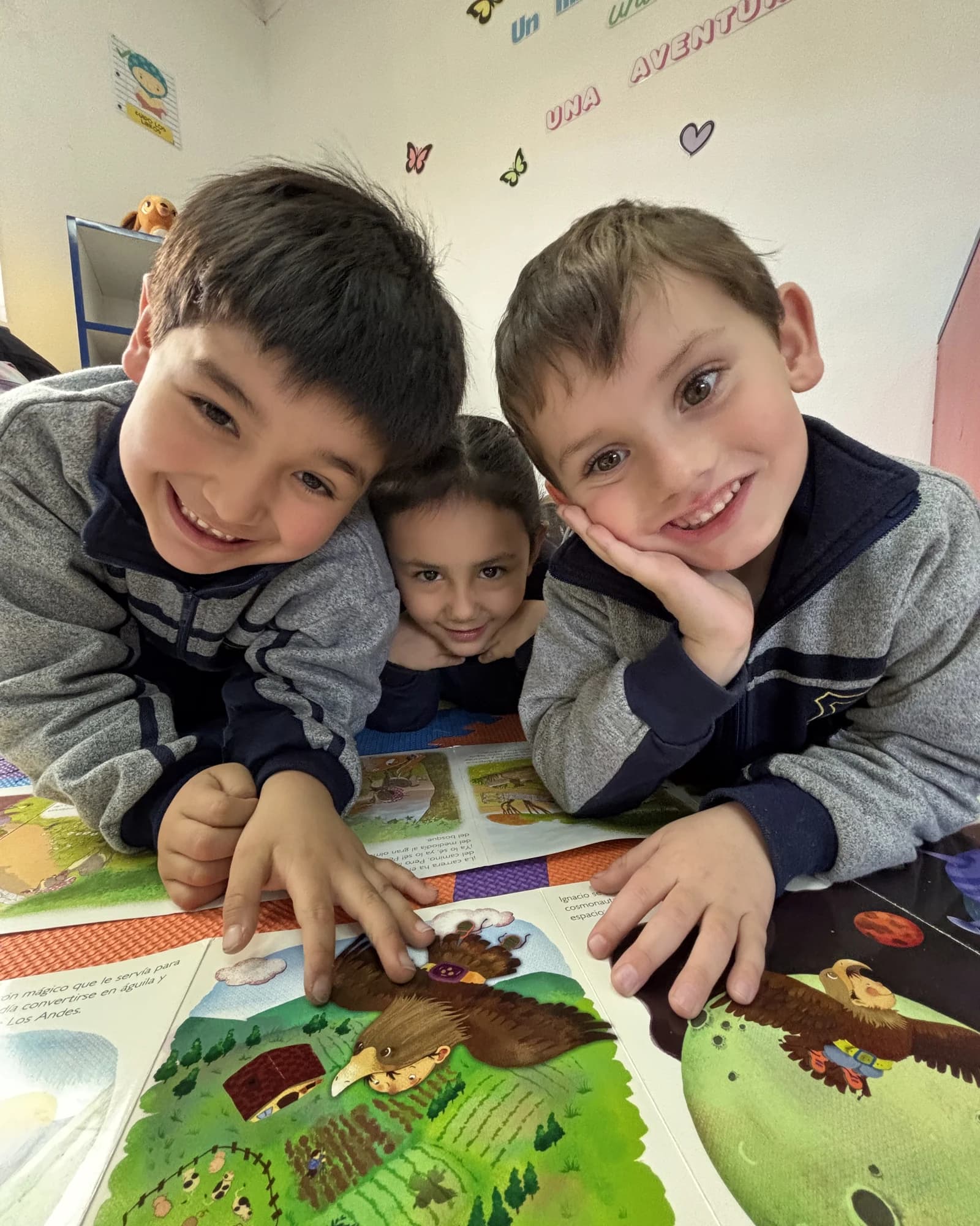 Tres párvulos del colegio leen juntos un libro ilustrado en la sala, bajo el letrero «Un libro, una aventura».