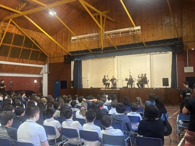 Gimnasio-teatro visto desde el fondo de la sala, con un grupo de músicos sobre el escenario y el público de estudiantes en las sillas azules.