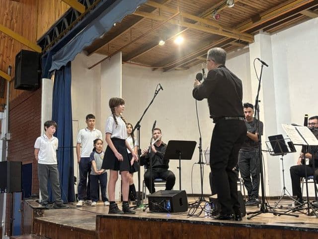 Presentación musical sobre el escenario del gimnasio-teatro: un grupo de estudiantes canta junto a un conjunto de músicos con atriles y micrófonos.