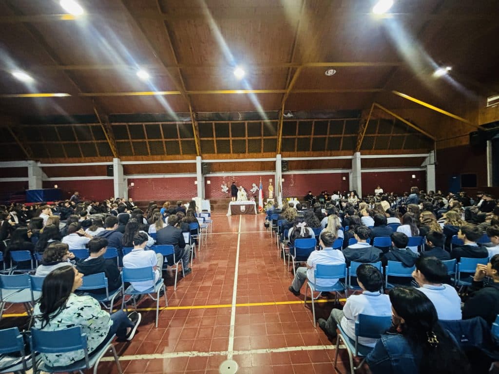 Gimnasio del colegio dispuesto como salón de actos, con dos bloques de sillas azules ocupados por toda la comunidad escolar y un altar al fondo.