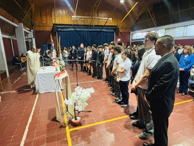 Misa en el gimnasio del colegio: el sacerdote junto al altar y los estudiantes de pie formando una fila a lo largo de la cancha, con las banderas al costado.