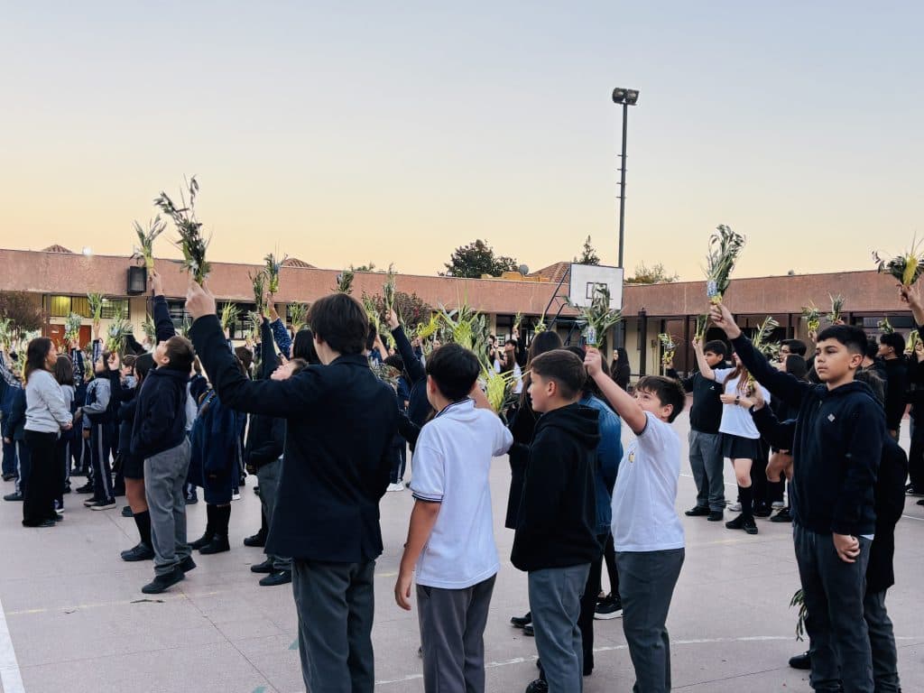 Celebración del Domingo de Ramos en el patio al atardecer: estudiantes de espaldas levantando ramos de palma trenzada frente a los edificios del colegio.