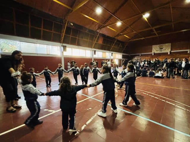 Estudiantes de los cursos menores forman una ronda tomados de la mano en el piso del gimnasio, con el estandarte del colegio colgado al fondo.