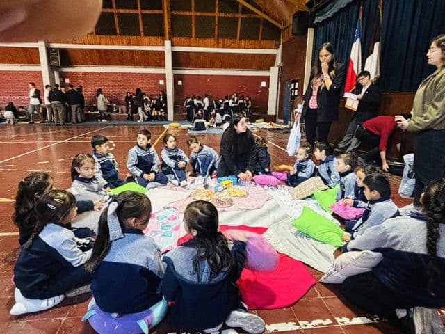 Estudiantes de los cursos menores sentados en círculo alrededor de un mantel con comida, acompañados por sus profesoras, en el gimnasio del colegio.