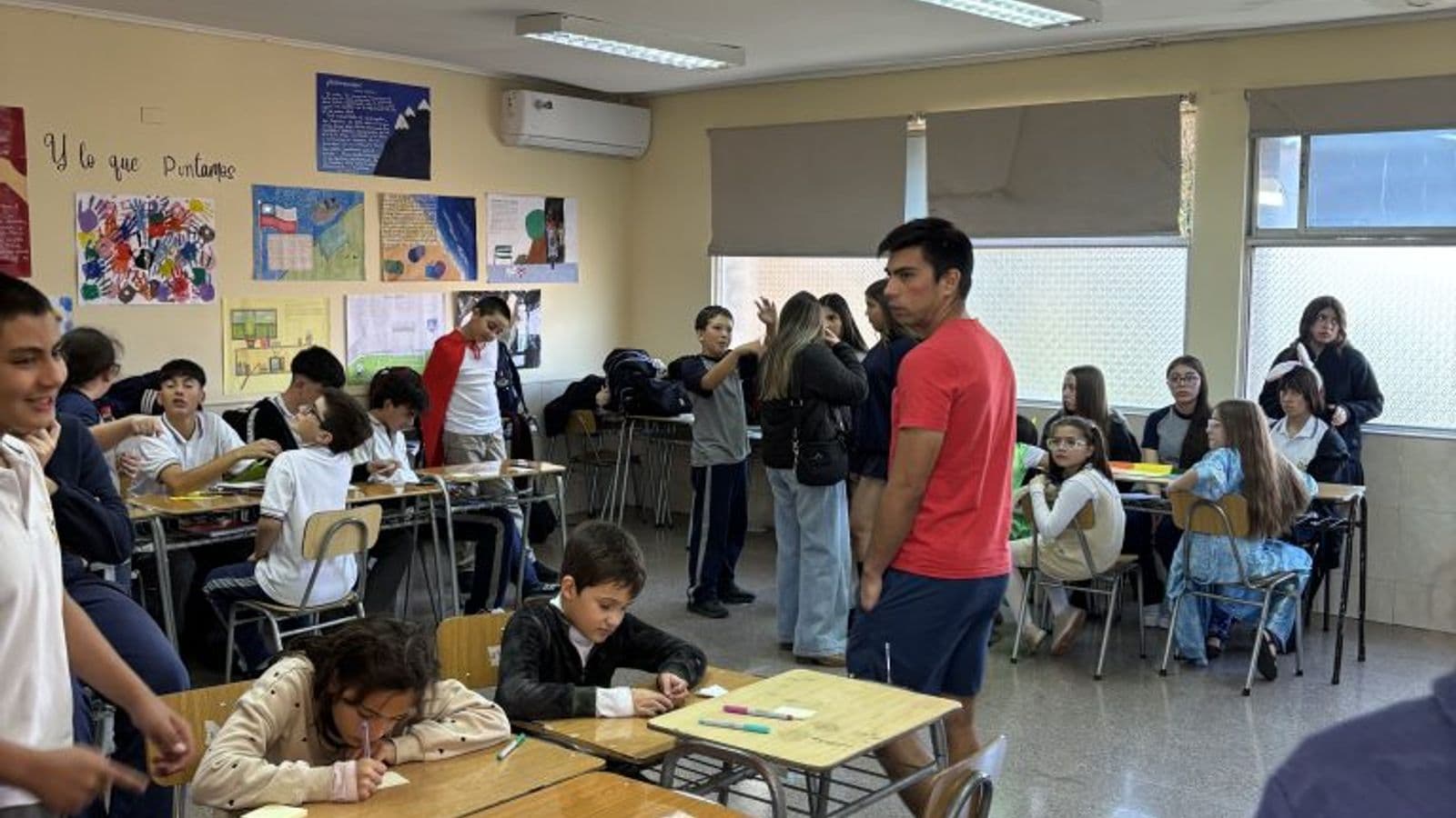 Sala de clases con los pupitres agrupados en islas para trabajo en equipo, murales de trabajos en la pared y estudiantes escribiendo.