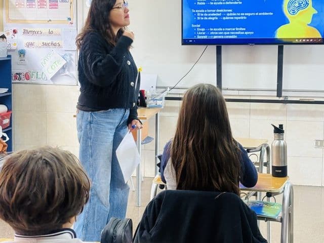 Clase sobre emociones: una profesional expone frente a una pantalla que explica para qué sirven las emociones, mientras los estudiantes escuchan desde sus puestos.