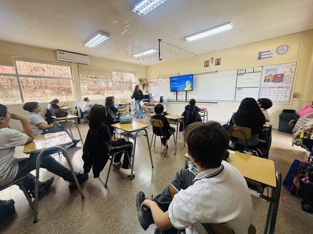 Sala de clases con ventanales a lo largo de un muro, pupitres individuales y una pantalla encendida; los estudiantes miran hacia el frente.