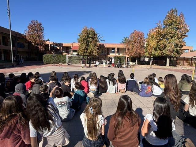 Patio central del colegio en otoño, con los edificios de dos pisos y los árboles al fondo bajo un cielo despejado; un grupo grande de estudiantes sentado en el suelo formando un círculo.
