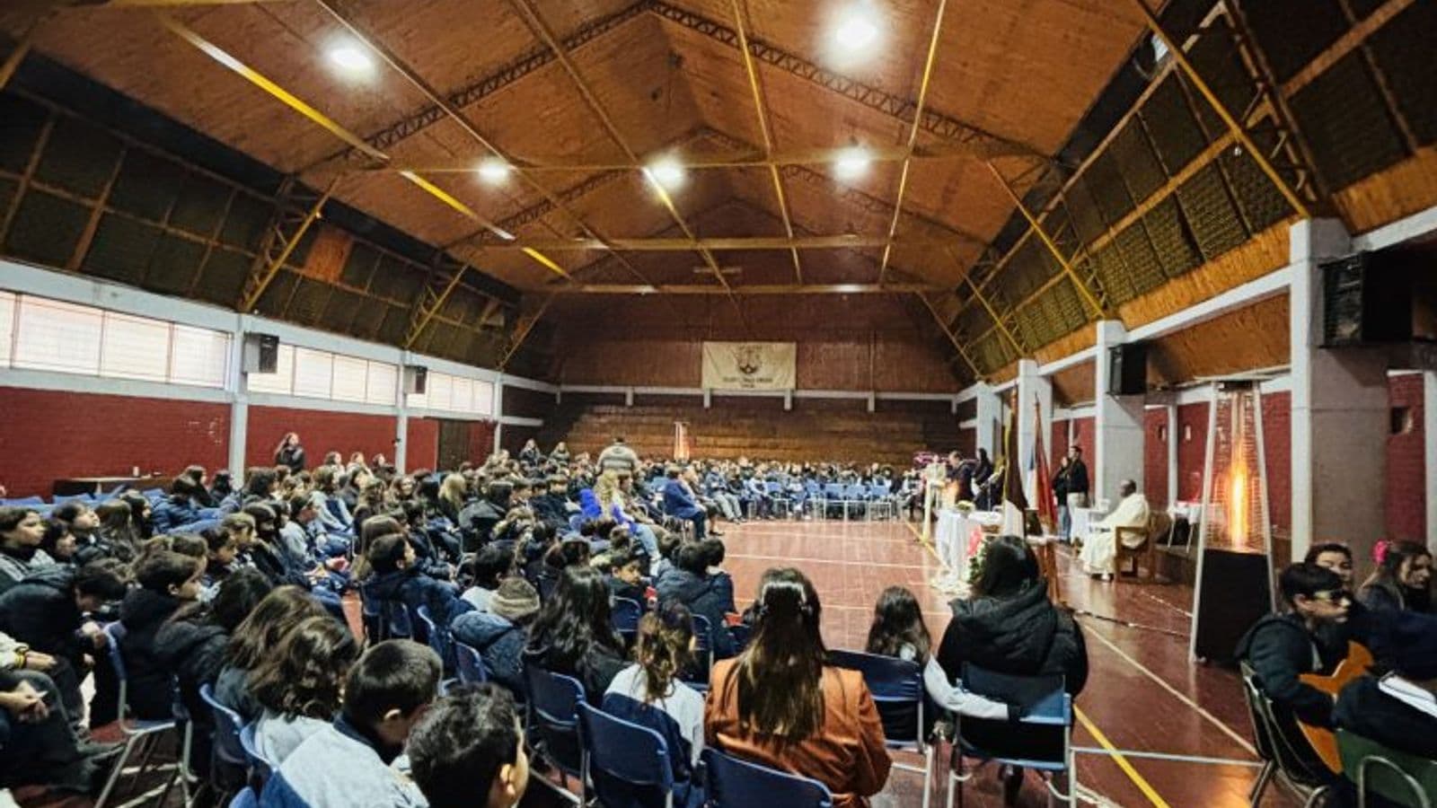 Interior del gimnasio-teatro en una celebración: techo de madera con luminarias, piso de baldosa roja con las líneas amarillas de la cancha y estudiantes sentados de espaldas.