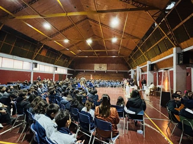 Interior del gimnasio-teatro en una celebración: techo de madera con luminarias, piso de baldosa roja con las líneas amarillas de la cancha y estudiantes sentados de espaldas.