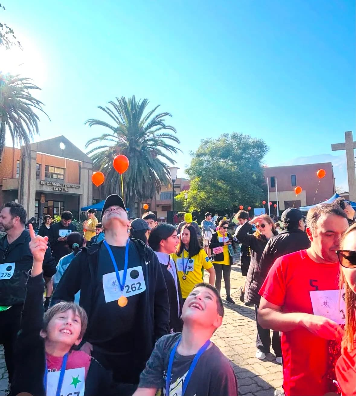 Familias y estudiantes con medallas frente a la fachada del colegio y la cruz del campus, en una kermesse con globos naranjos.