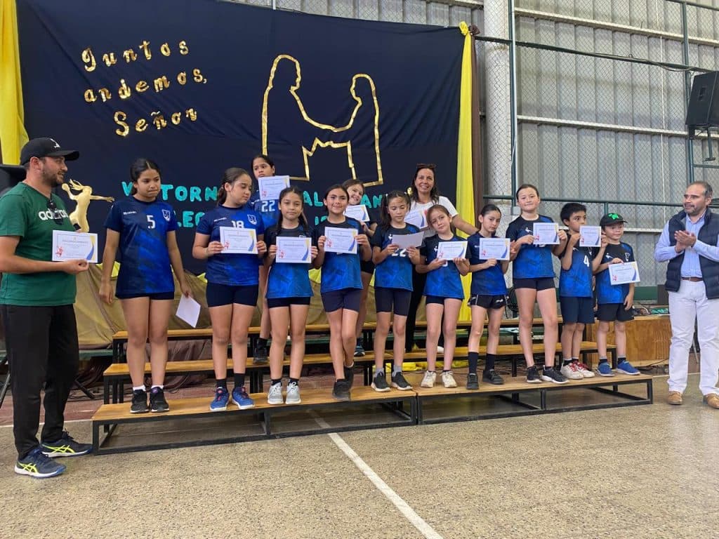 Premiación deportiva en el gimnasio: un equipo de estudiantes con su uniforme de juego recibe diplomas sobre una tarima, frente a un lienzo que dice «Juntos andemos, Señor».