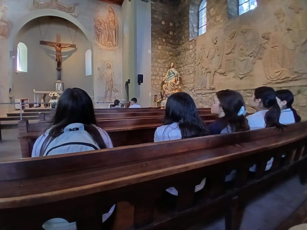 Estudiantes sentados de espaldas en las bancas de una iglesia de piedra, frente al altar y a un gran relieve tallado en el muro.