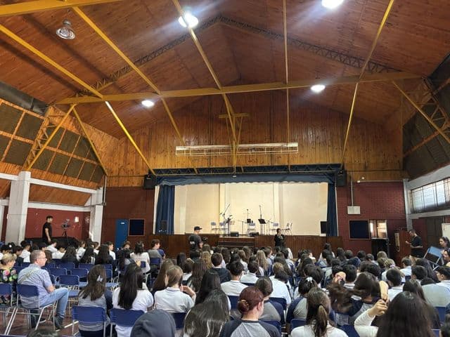 Vista general del gimnasio-teatro durante una actividad: hileras de sillas azules ocupadas por estudiantes y el escenario iluminado al fondo.