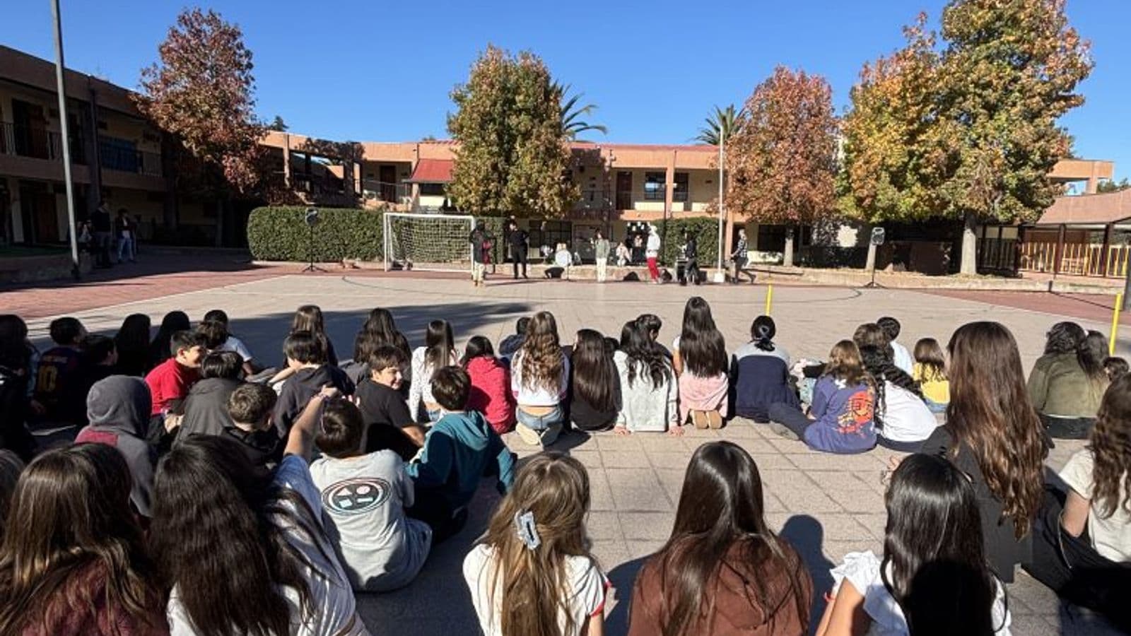 Patio central del colegio en otoño, con los edificios de dos pisos y los árboles al fondo bajo un cielo despejado; un grupo grande de estudiantes sentado en el suelo formando un círculo.